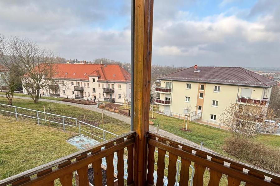 Blick von einem Holzbalkon auf ein gegenüberliegends mehrstöckickes Wohnhaus, dahinter eine entfernte Stadtansicht.