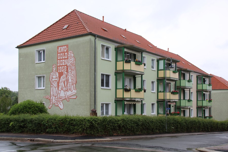 Mehrstöckige Wohnhäuser mit hellgüner Fassade, rotem Satteldach, Fenstern und Balkonen. Davor eine grüne Hecke an einer Straße.