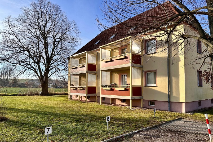 Mehrstöckiges Wohnhaus mit gelber Fassade, rotem Satteldach, Fenstern und Balkonen und einer Grünfläche und PKW-Stellflächen davor.