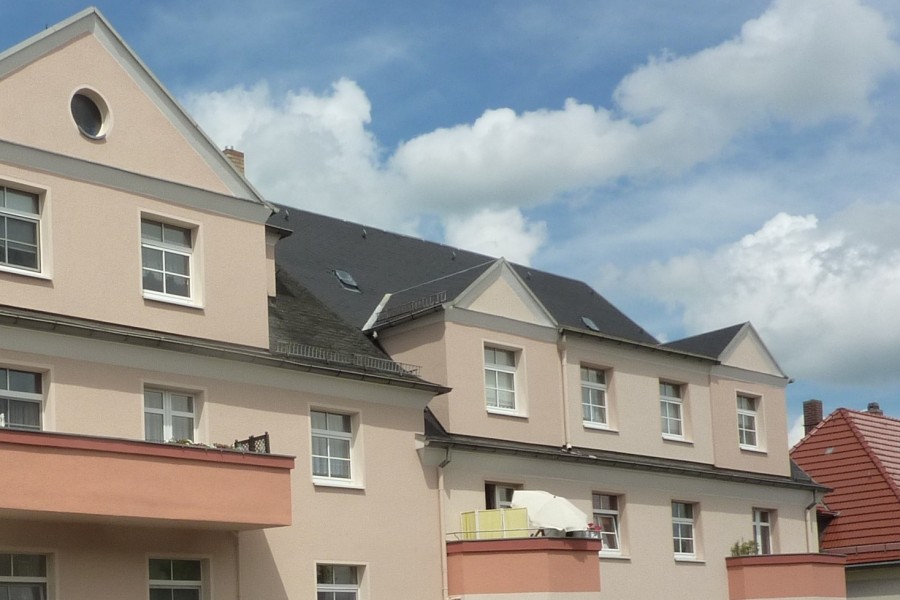 Mehrstöckiges Wohnhaus mit heller Fassade, dunkelgrauem Satteldach und Fenstern und Balkonen. Davor ein weißer Gartenzaun.