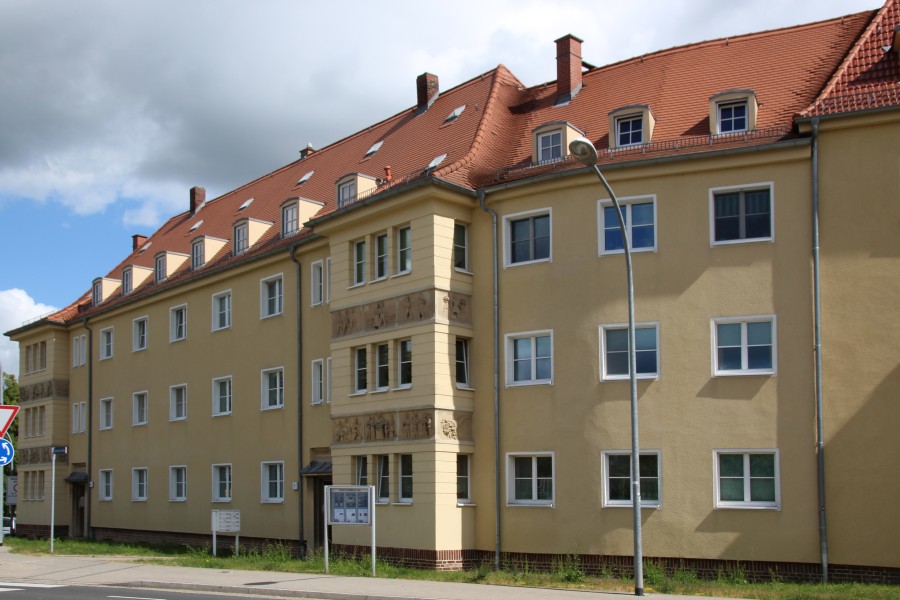 Mehrstöckiges Wohnhaus mit gelber Fassade, rotem Satteldach und Fenstern an einer Straße.