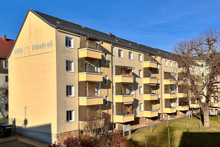 Mehrstöckiges Wohnhaus mit gelber Fassade, dunkelgrauem Dach, Fenstern und Balkonen, davor Grünfläche mit Wäscheplatz, Hecken und einem Baum.