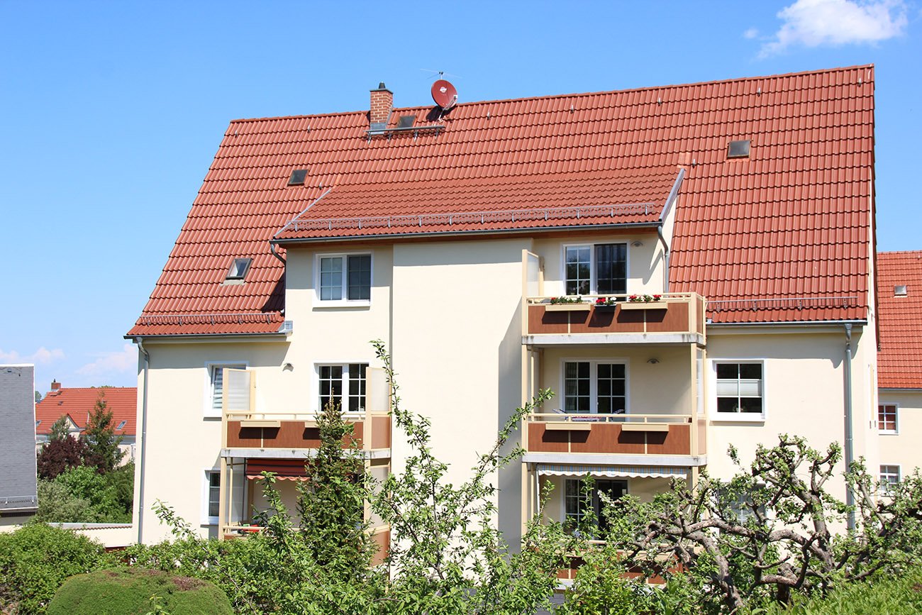 Zweistückiges Mehrfamilienhaus mit Balkonen, blauer Himmel und davor kleine Bäume und eine Hecke.