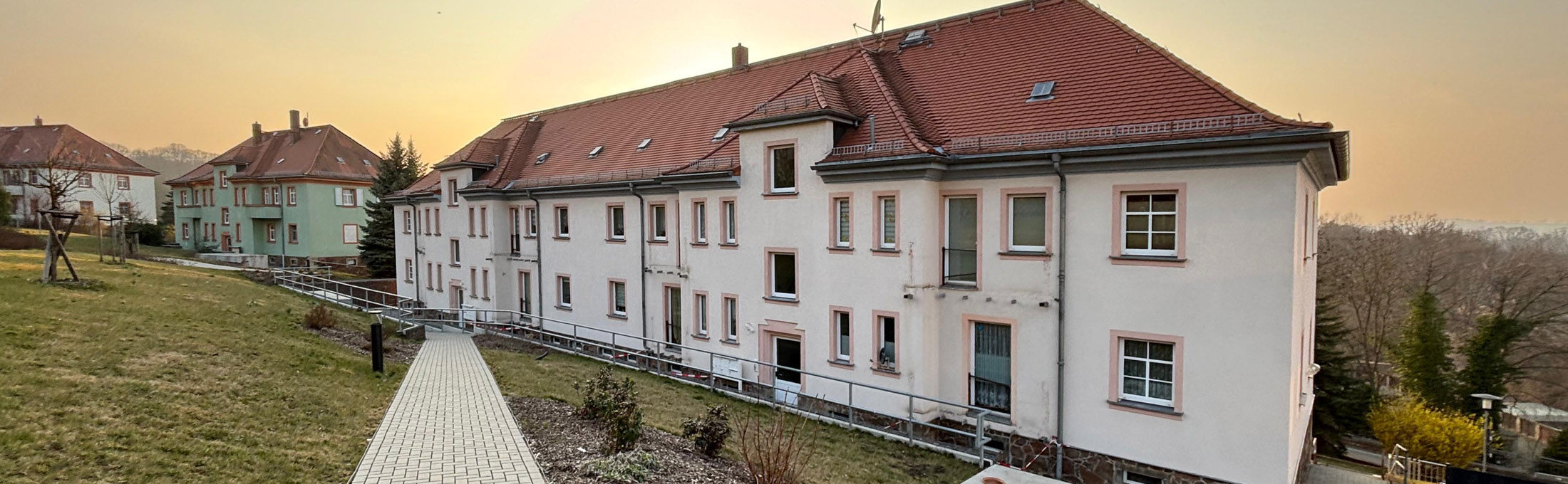 Mehrere zweistöckige Mehrfamilienhäuser mit einem gepflastertem FUßweg davor und Grünfläche zur Sonnenuntergangszeit.