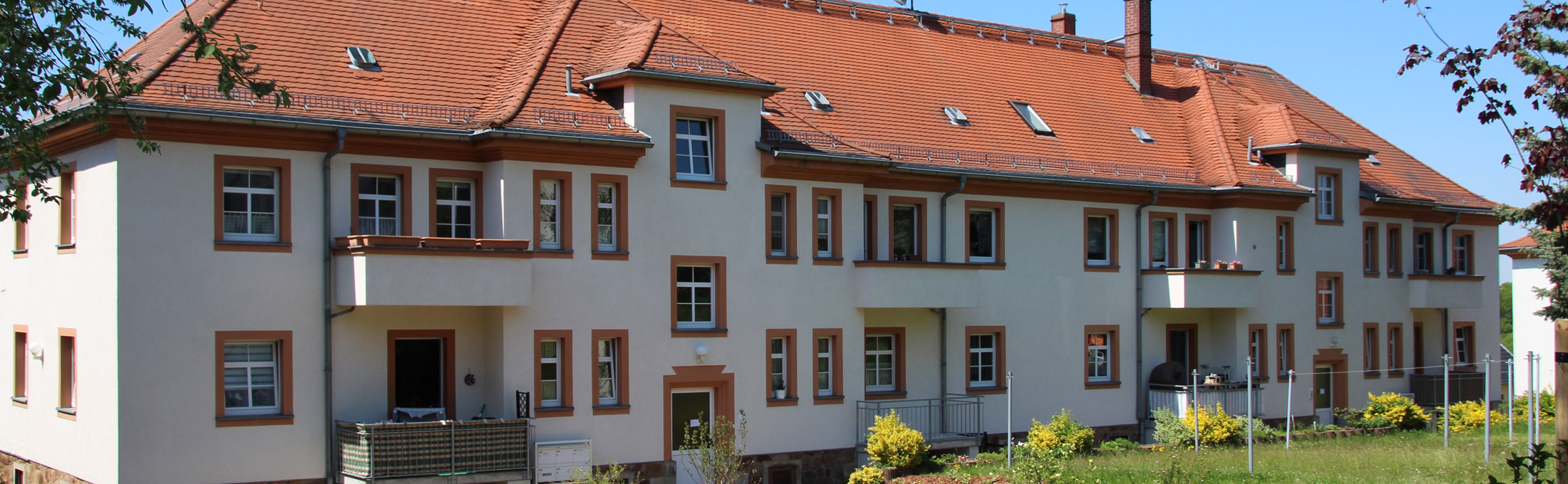 Zweistöckiges Mehrfamilienhaus mit einer Grünfläche und blühenden Forsythien-Sträuchern davor.